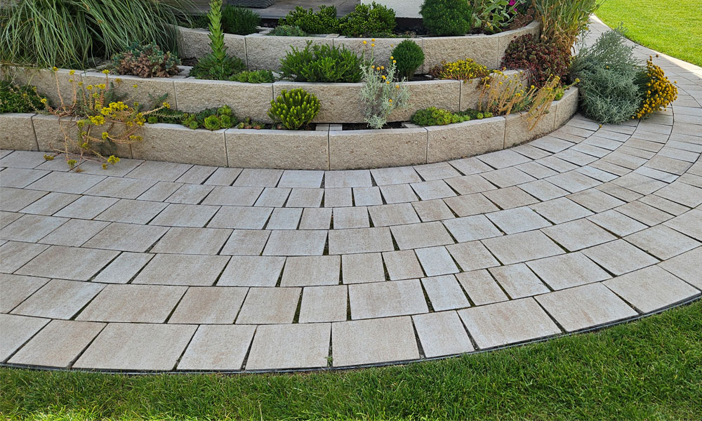 A Friedl Steinwerke Piazza mészkő árnyalt színben, íves lerakásban, egy teraszos, növényekkel beültetett rész mellett. A Friedl Steinwerke Piazza mészkő árnyalt színben, íves lerakásban, egy teraszos, növényekkel beültetett rész mellett.